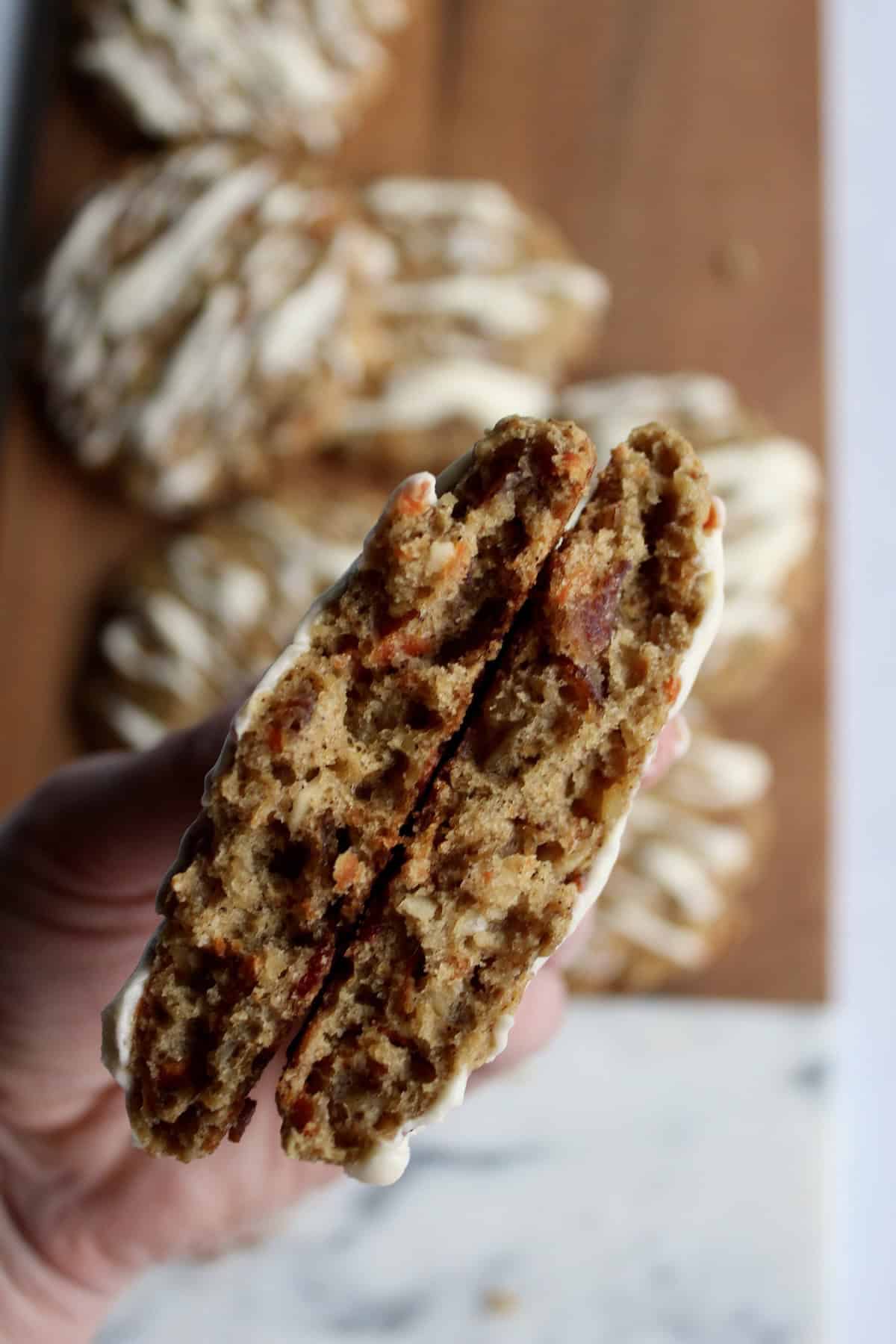 an oatmeal carrot cake cookie is broken in half and held to show the soft texture and chunky mix-ins