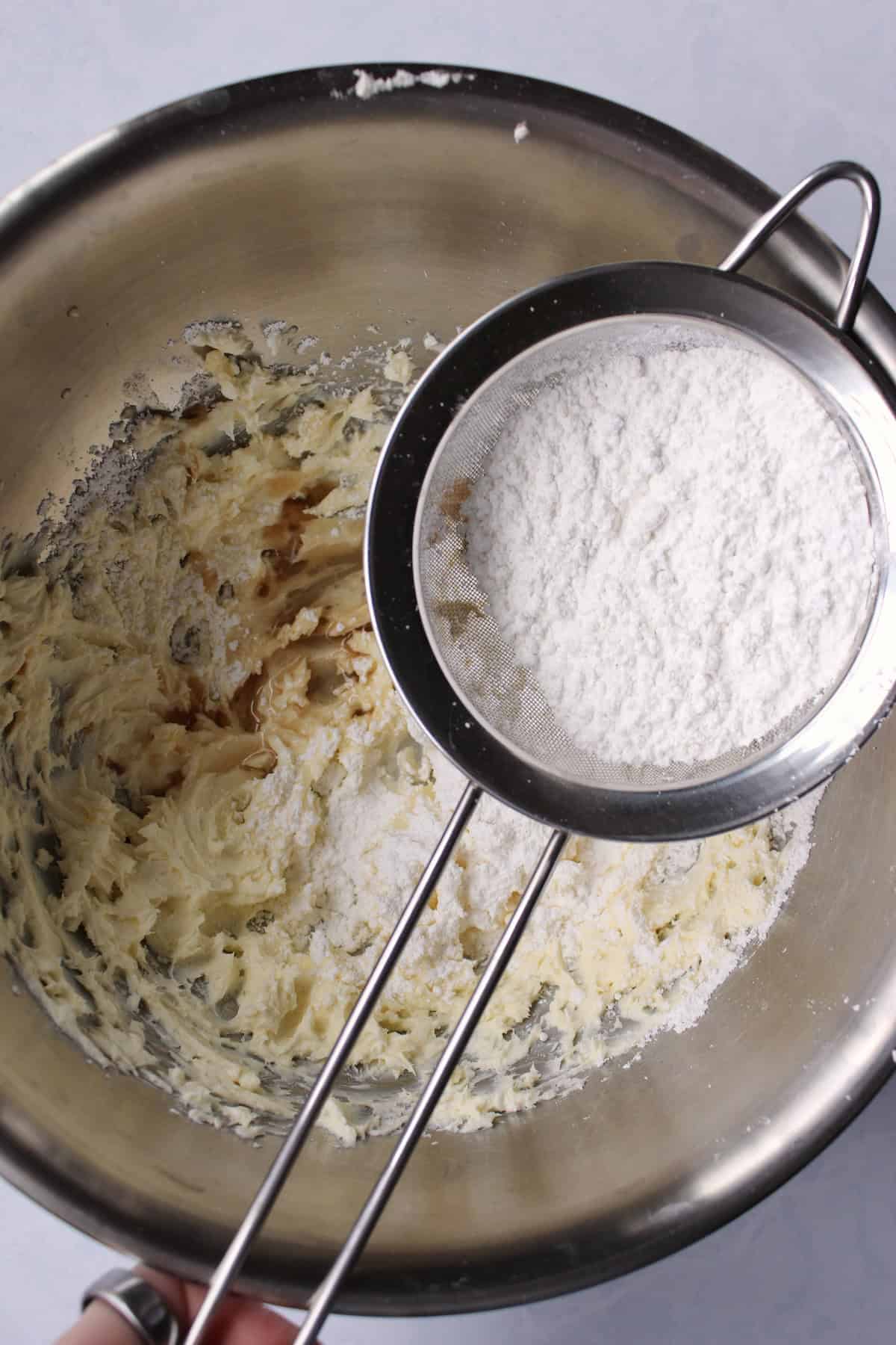 a sifter of powdered sugar rests over the surface of the bowl with the butter and cream cheese mixture