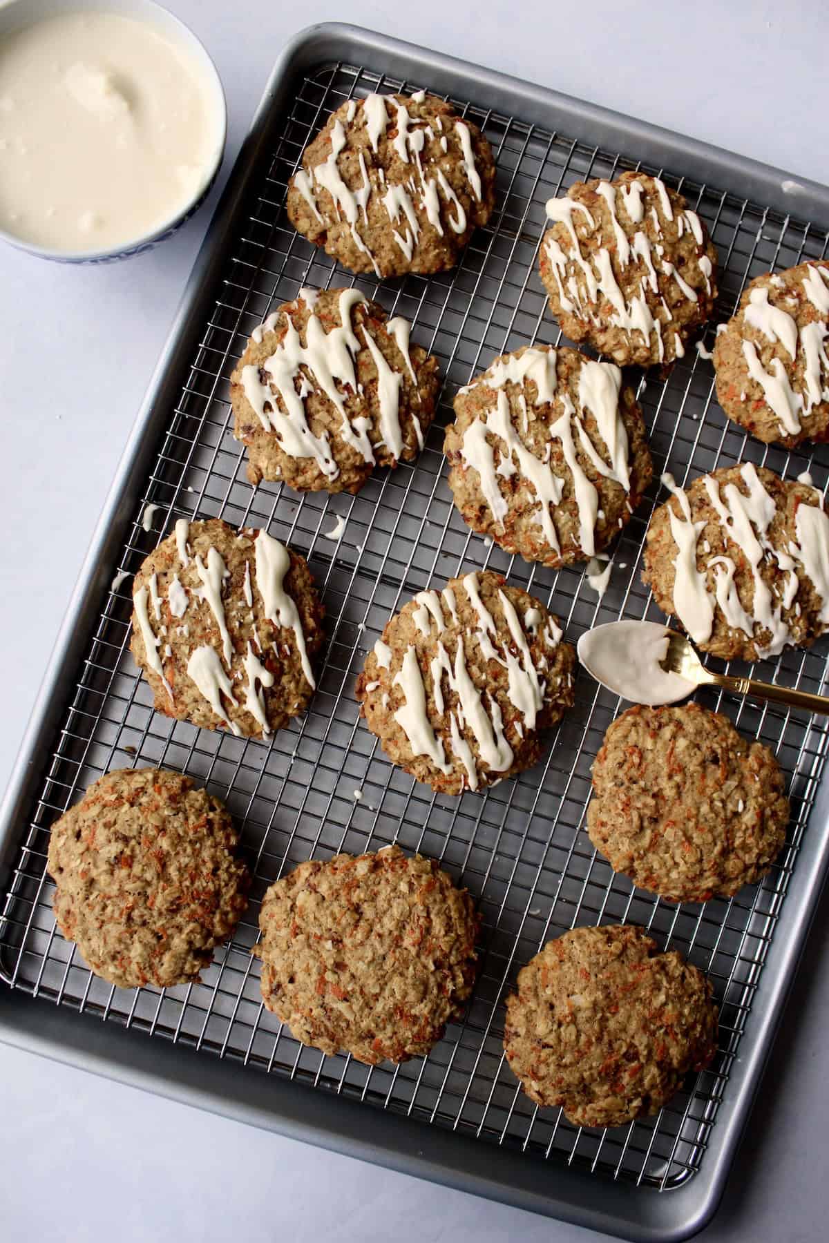 a small metal spoon is used to drizzle the cream cheese glaze over the baked carrot cake oatmeal cookies, creating diagonal lines and speckles