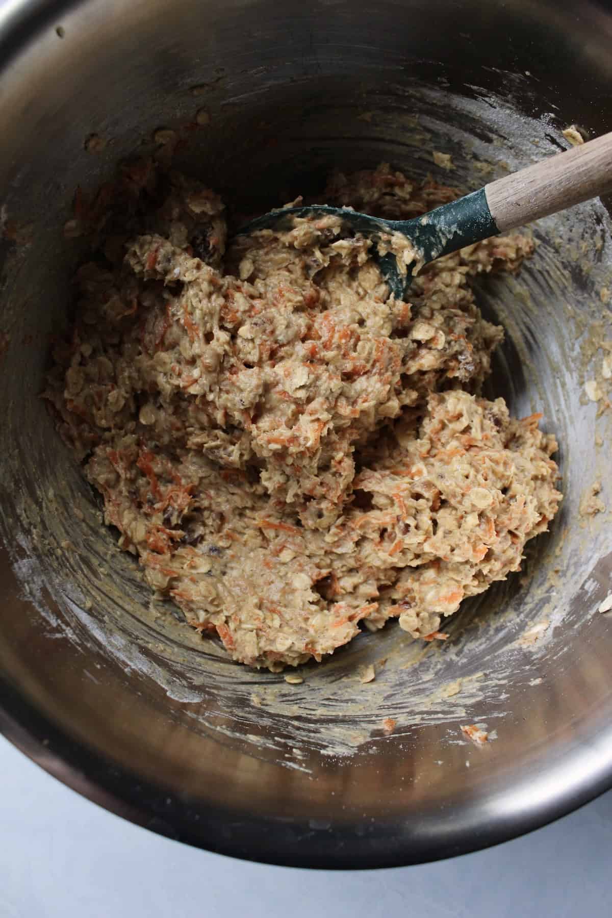 a chunky, thick dough comes together in a large mixing bowl, with visible shreds of carrots, oats, and pieces of nut