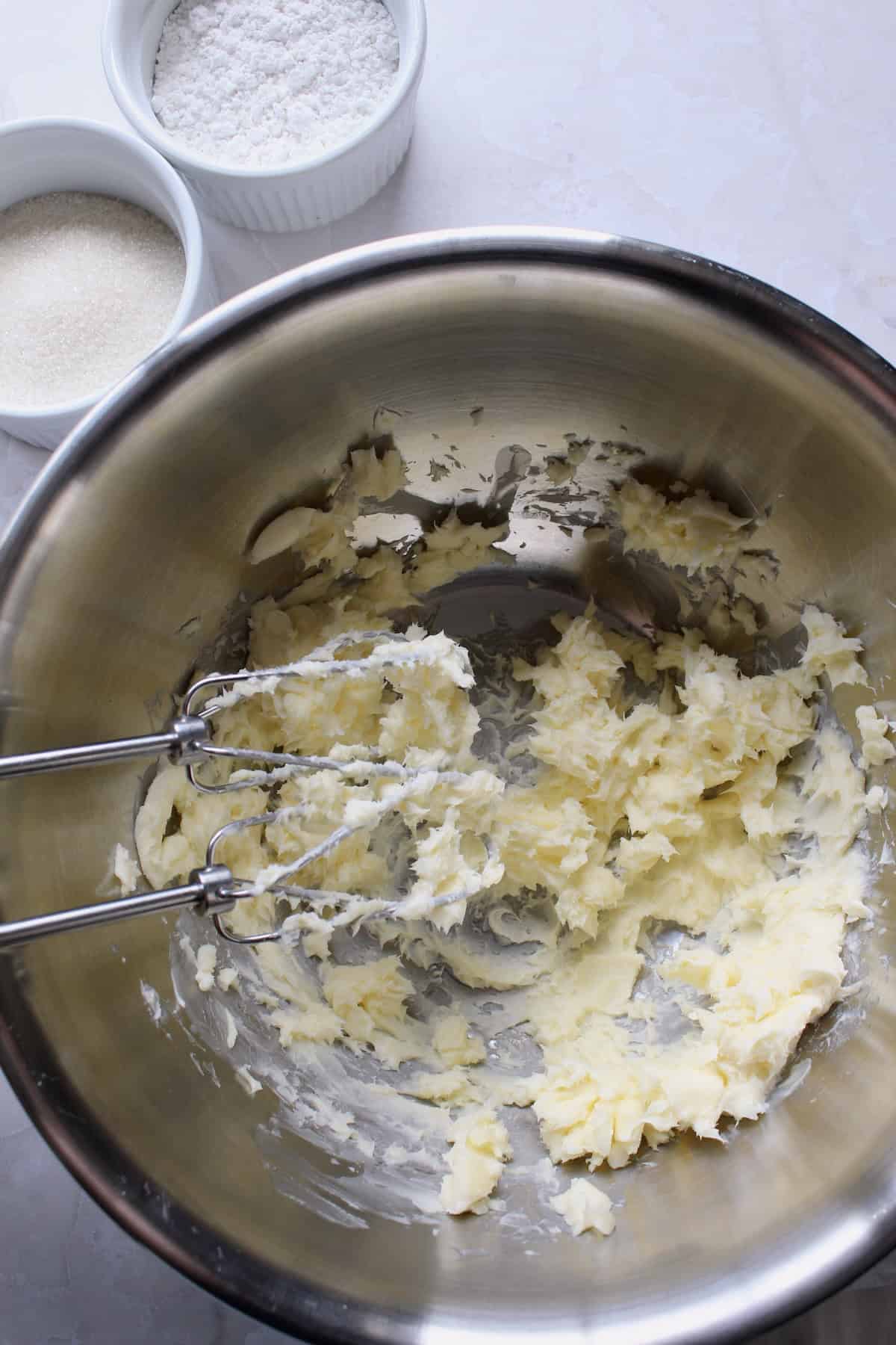 butter is whipped in a large mixing bowl next to two smaller bowls of powdered sugar and granulated sugar