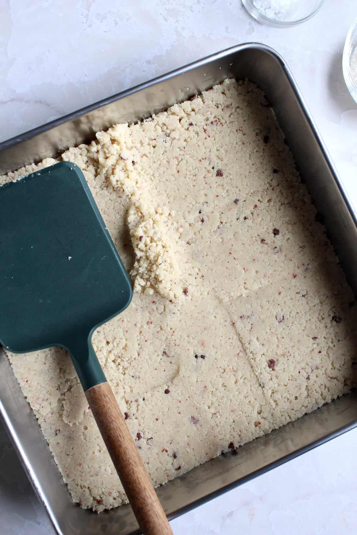 a wide spatula is used to press the surface of the shortbread cookie dough, flattening it into the baking pan and creating a smooth surface