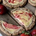 a halved strawberry cheesecake cookie is set on a wooden board along with several whole cookies and a few fresh strawberries