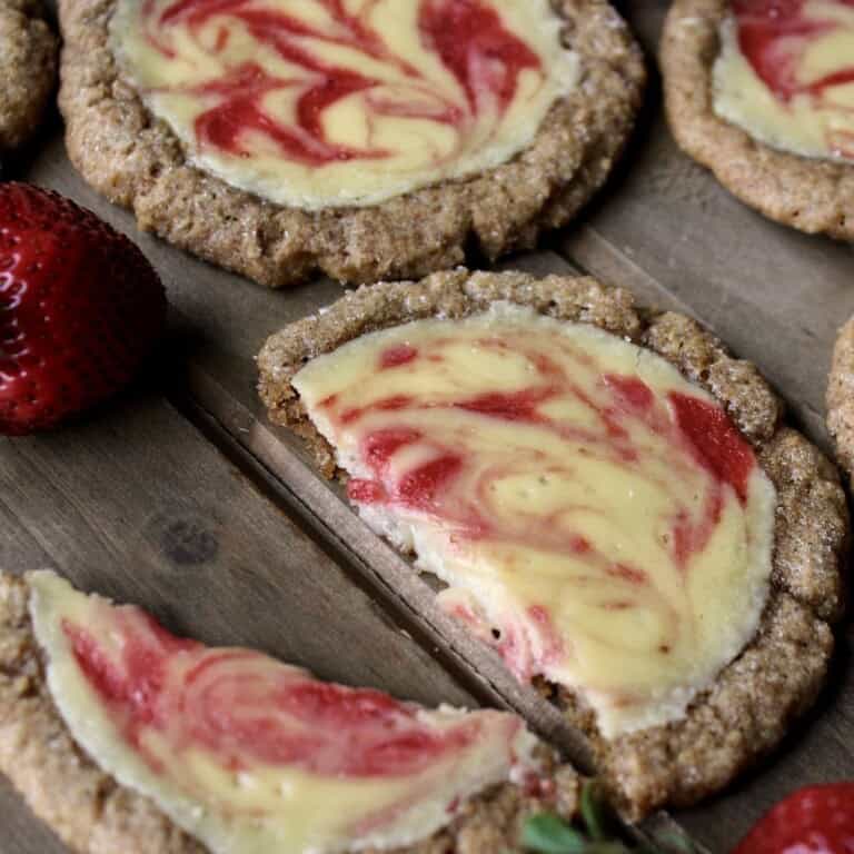 a halved strawberry cheesecake cookie is set on a wooden board along with several whole cookies and a few fresh strawberries