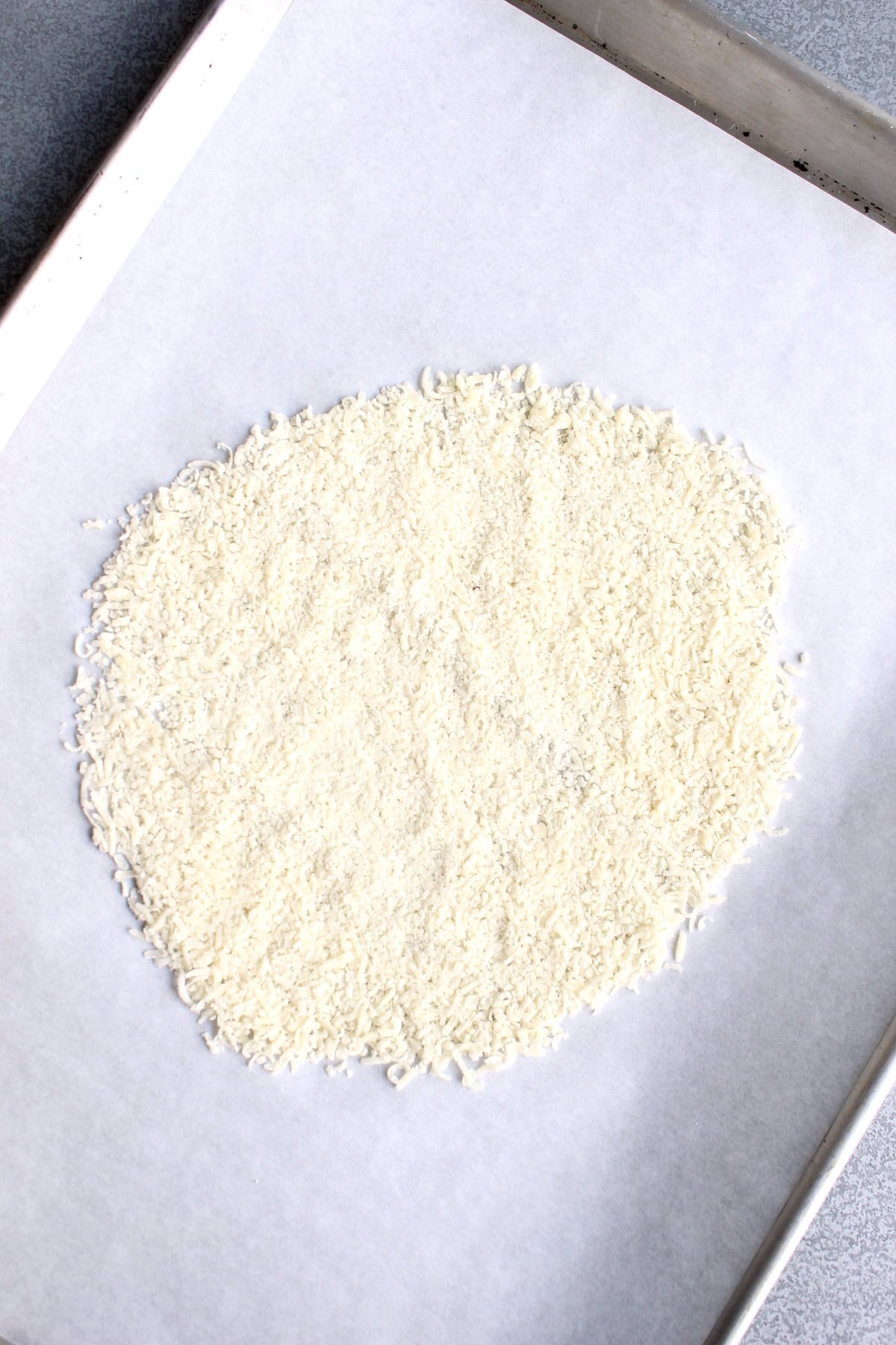 freshly grated parmesan cheese is smoothed into a flat disc on a parchment paper lined baking sheet.