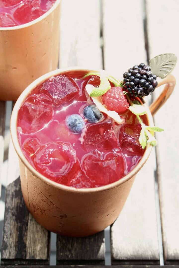 a berry moscow mule drink sits on a wooden patio table, with the ice sparkling in the sun