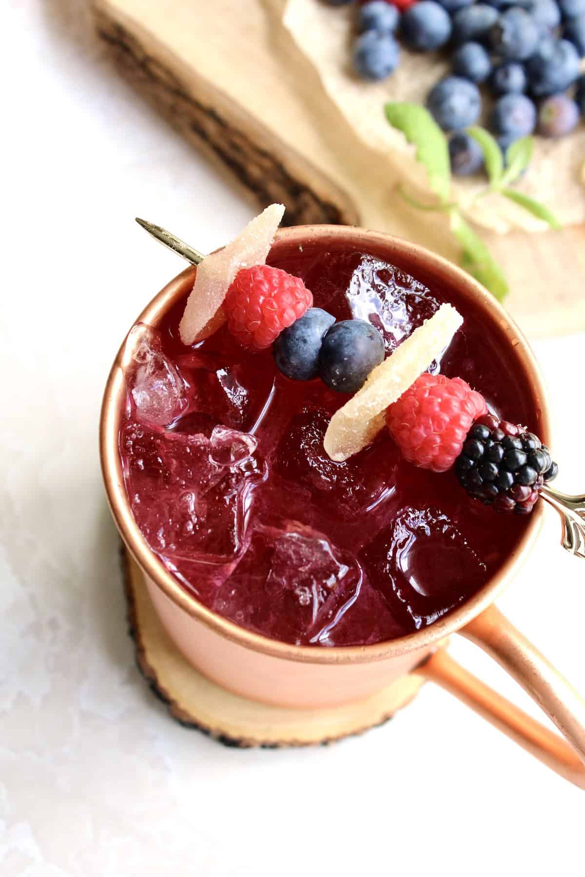 a berry moscow mule cocktail is garnished with a skewer that has alternating types of berries and small slices of candied ginger