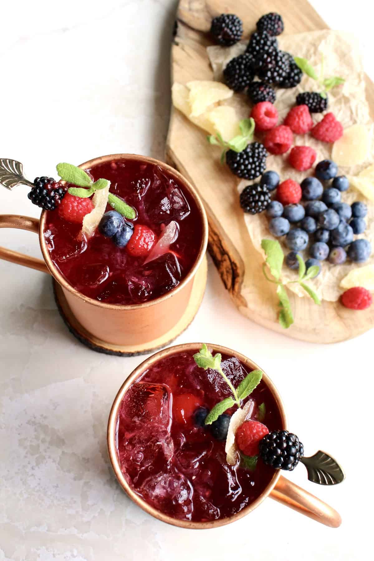 two berry moscow mules are garnished with skewers of fresh berries and candied ginger, along with fresh mint sprigs.