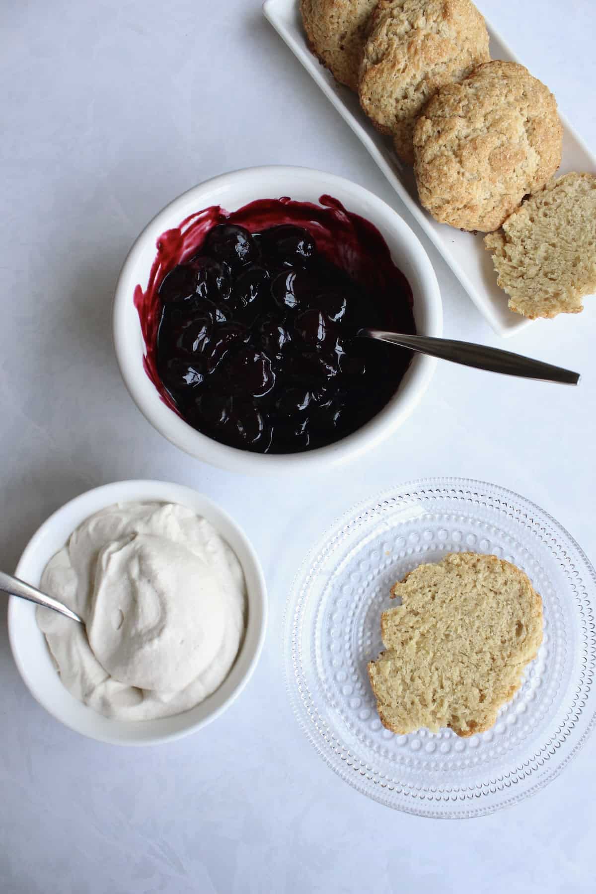 the bottom half of a shortcake biscuit rests on a glass plate near bowls of cherry compote and whipped cream