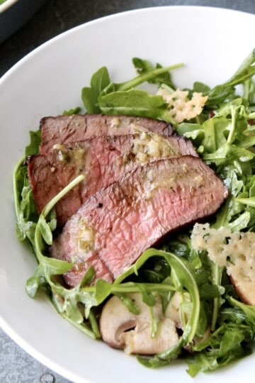 arugula steak salad with slices of medium rare steak and parmesan crisps is served in a white salad bowl