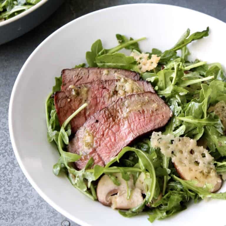arugula steak salad with slices of medium rare steak and parmesan crisps is served in a white salad bowl