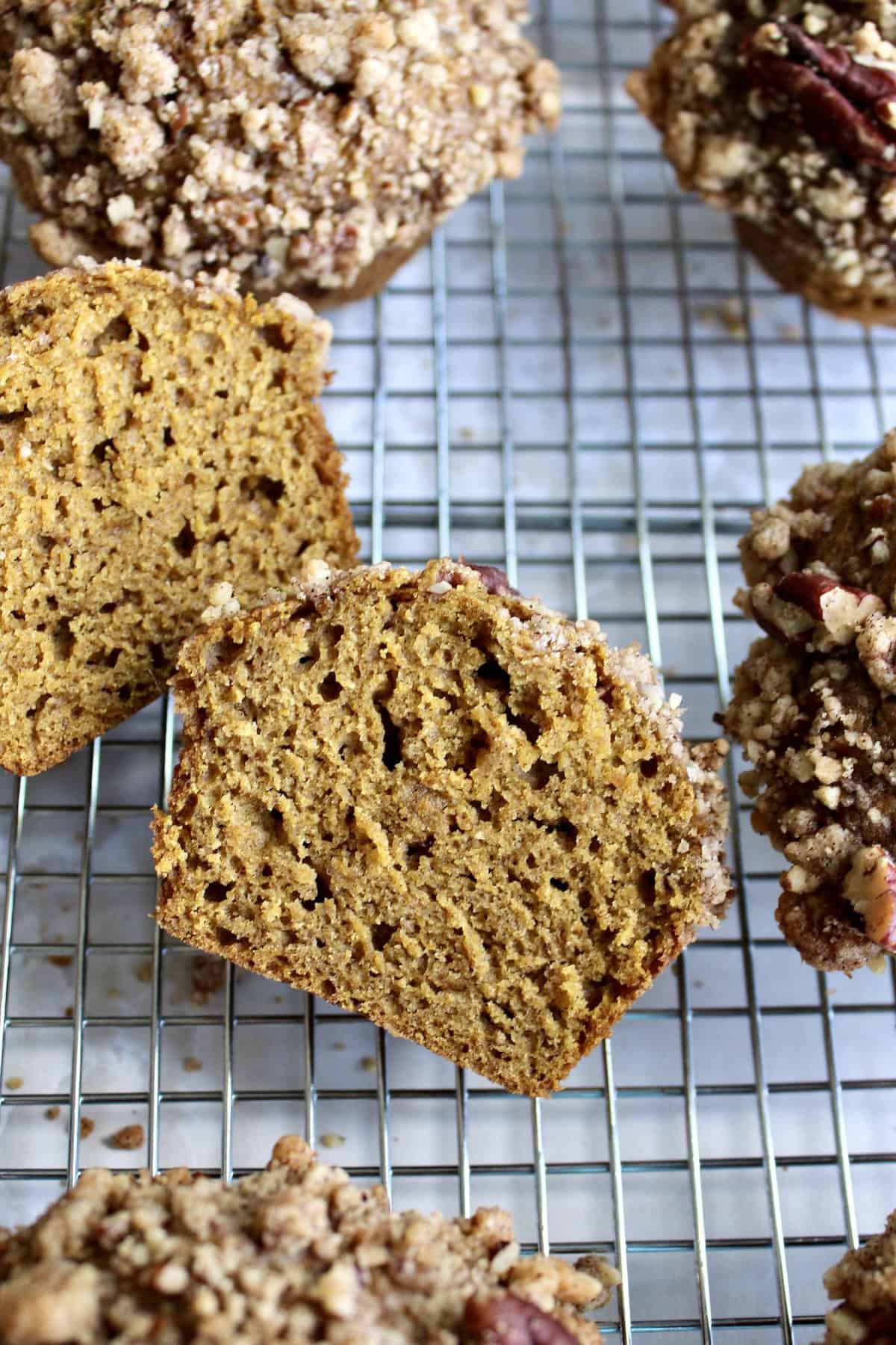 two halves of a pumpkin muffin with pecan streusel surrounded by whole muffins on a wire cooling rack