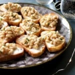 a platter of white bean bruschetta on a table with gold rimmed wine glasses and twinkle lights