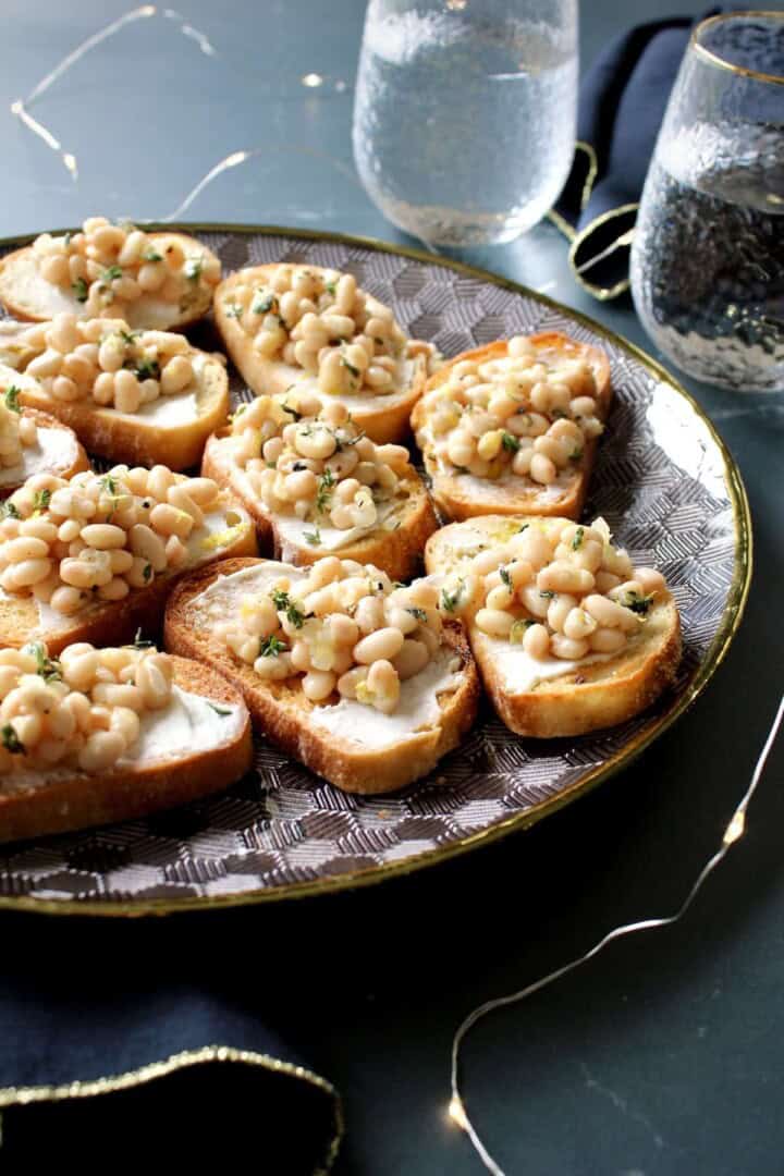 a platter of white bean bruschetta on a table with gold rimmed wine glasses and twinkle lights