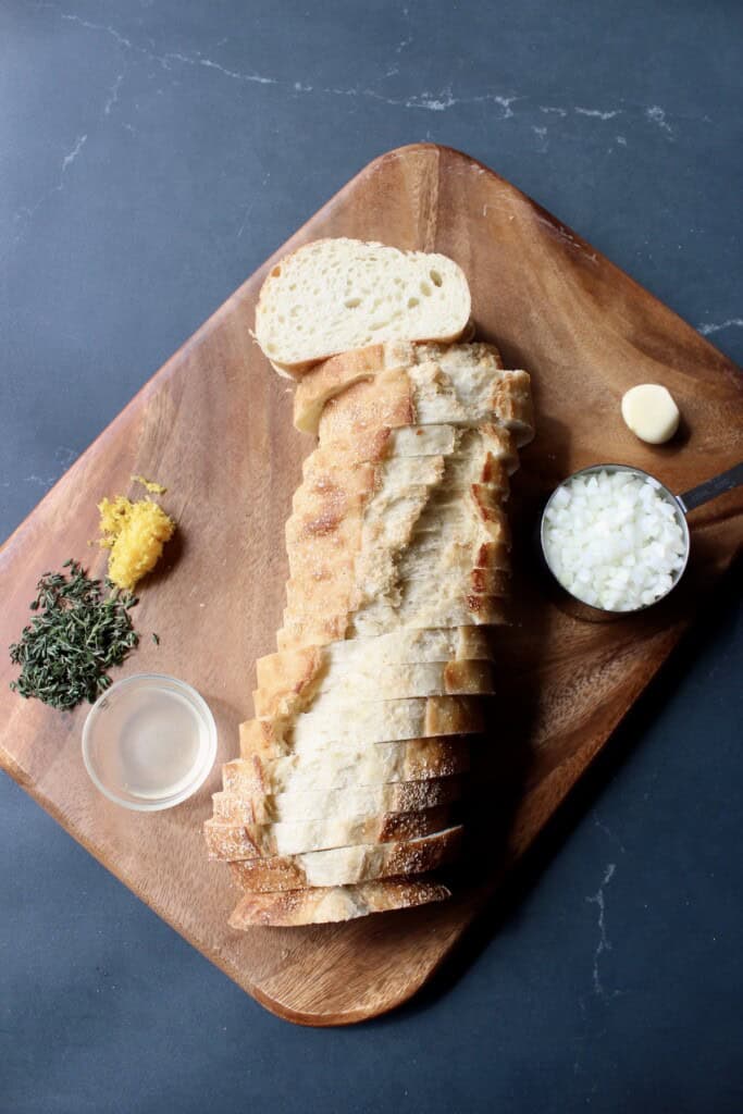 sliced bread, chopped onion, fresh thyme, and garlic are prepped on a wooden cutting board.